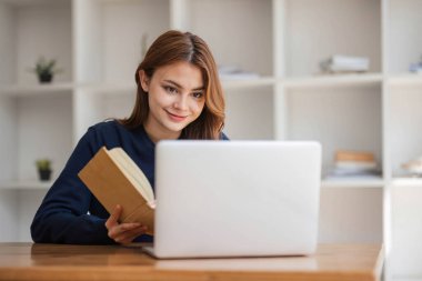 Asyalı üniversite öğrencisi kız öğrenci kütüphanede dizüstü bilgisayarlarıyla ödev için araştırma yapıyor, ödev ödevi için notlar alıyor, online eğitim e-öğrenim kavramı