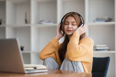 Kulaklıklı Asyalı bir kadın internetten ders çalışmaya ara verip iş sırasında müzik dinlemekten mutlu bir şekilde müzik dinliyor.