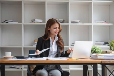 Resmi takım elbiseli Asyalı iş kadını bilgisayarını kullanıyor ve ofisindeki masasında pazarlama raporları üzerinde çalışıyor.