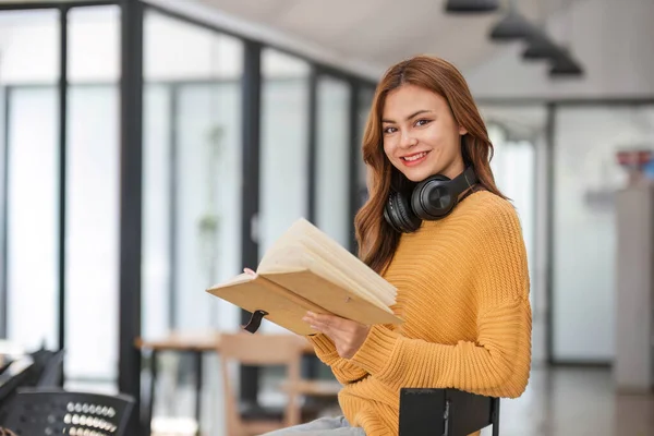 stock image A portrait of an attractive young Asian female in a cozy yellow sweater stands inside a coffee shop with a book and headphones. college student, freelancer..