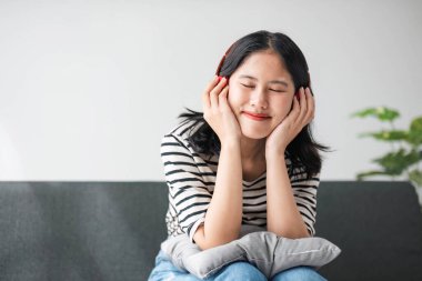 Kanepede müzik dinlemek için kulaklıklı akıllı cep telefonu kullanan Asyalı genç bir kadın portresi...