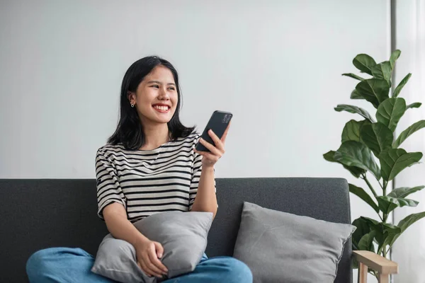 stock image Asian woman using smartphone while laying on the sofa at home. Cheerful lady enjoys online chatting, spends time in social networks, looks at the phone and smiles..