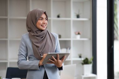 Asyalı başarılı bir Müslüman iş kadınının portresi modern ofisinde elinde dijital tabletle duruyor....