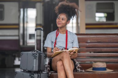 Asyalı Afrikalı bayan turist elinde cep telefonuyla tren istasyonunda oturuyor. İstasyonda akıllı telefon dizüstü bilgisayarı oynayan kendine güvenen genç kız.
