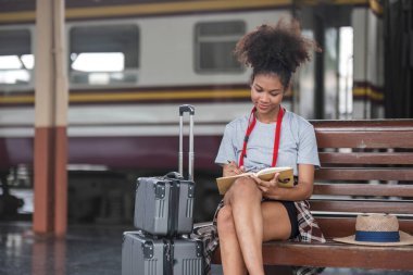 Asyalı Afrikalı bayan turist elinde cep telefonuyla tren istasyonunda oturuyor. İstasyonda akıllı telefon dizüstü bilgisayarı oynayan kendine güvenen genç kız.