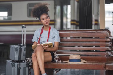 Asyalı Afrikalı bayan turist elinde cep telefonuyla tren istasyonunda oturuyor. İstasyonda akıllı telefon dizüstü bilgisayarı oynayan kendine güvenen genç kız.