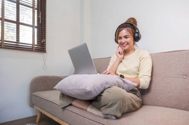 Genç ve güzel Asyalı kadın, dinleniyor ve kulaklıkla müzik dinliyor. Kanepede yatıyor. Genç bir kadın evde dizüstü bilgisayar kullanıyor....