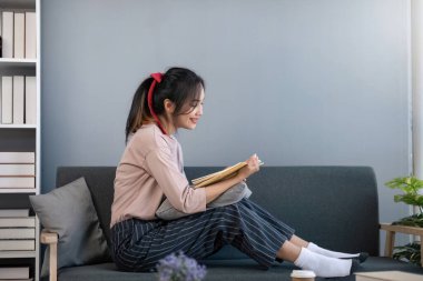 Gülümseyen Asyalı kadın evde kitap okuyor, kanepede dinleniyor..