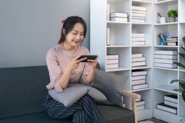 Cep telefonu kullanan mutlu Asyalı kadın. Akıllı telefon kullanan gülümseyen genç bir kadın. Sosyal medyada oyna. Evdeki kanepede rahatla...