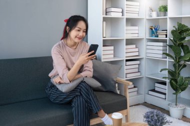 Cep telefonu kullanan mutlu Asyalı kadın. Akıllı telefon kullanan gülümseyen genç bir kadın. Sosyal medyada oyna. Evdeki kanepede rahatla...