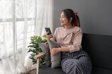Cep telefonu kullanan mutlu Asyalı kadın. Akıllı telefon kullanan gülümseyen genç bir kadın. Sosyal medyada oyna. Evdeki kanepede rahatla...