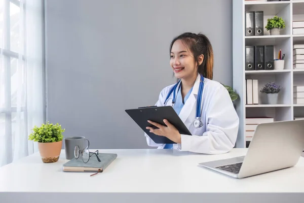 stock image Asian doctor young beautiful woman smiling using working with a laptop computer and her writing something on paperwork or clipboard white paper at hospital desk office, Healthcare medical concept.