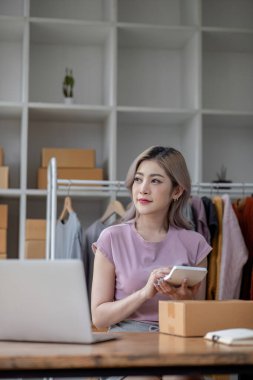 Dişi çevrimiçi satıcı teslimat ögelerini hazırlıyor ve siparişleri hesaplamak için hesap makinesi kullanıyor.