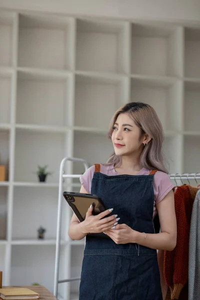 businesswoman owner an online trading business is using tablet to check the stock of the clothes on the rail rack at small office at home. work from home, online delivery, SME..