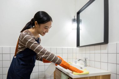 Genç bir hizmetçi bez ve temizlik spreyi kullanarak banyoyu temizliyor...