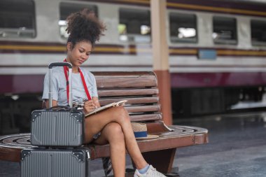 Asyalı Afrikalı bayan turist elinde cep telefonuyla tren istasyonunda oturuyor. İstasyonda akıllı telefon dizüstü bilgisayarı oynayan kendine güvenen genç kız.