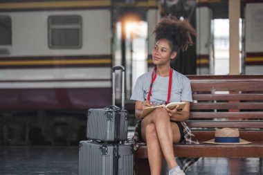 Asyalı Afrikalı bayan turist elinde cep telefonuyla tren istasyonunda oturuyor. İstasyonda akıllı telefon dizüstü bilgisayarı oynayan kendine güvenen genç kız.