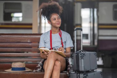 Asyalı Afrikalı bayan turist elinde cep telefonuyla tren istasyonunda oturuyor. İstasyonda akıllı telefon dizüstü bilgisayarı oynayan kendine güvenen genç kız.