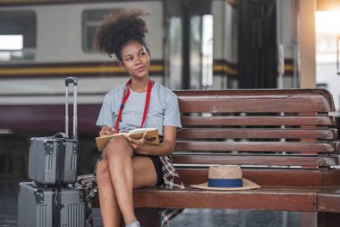 Asyalı Afrikalı bayan turist elinde cep telefonuyla tren istasyonunda oturuyor. İstasyonda akıllı telefon dizüstü bilgisayarı oynayan kendine güvenen genç kız.