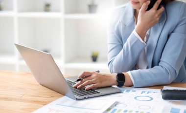 İnternetteki toplantıda dizüstü bilgisayar kullanan çekici Asyalı kadın. İş kadını cep telefonu kullanıyor. Modern ofiste masada oturan iş arkadaşıyla konuşuyor..