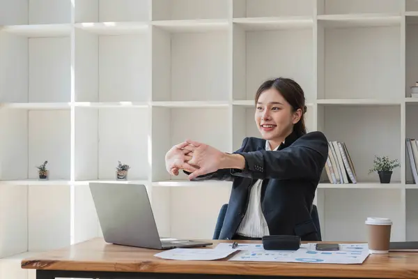A happy and beautiful Asian businesswoman stretched her arms after finishing her work on a laptop at her desk, feeling carefree and comfortable at her workplace..