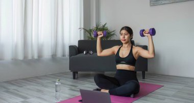 Dizüstü bilgisayarın önünde egzersiz yapan genç bir kadın spor barı kıyafeti giyer. Yoga yap ve egzersiz paspasına halterleri kaldır...