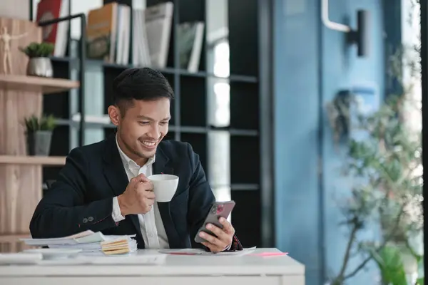 Asian businessman office worker drinking coffee and having fun chatting on a video call on a smartphone with friends during lunch break