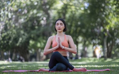 30 'lu yaşlarındaki çekici ve çekici Asyalı kadın spor kıyafetleriyle yoga yapıyor, kaslarını gevşetiyor. Güzel yeşil bir parkta yoga yapan Asyalı kadın..