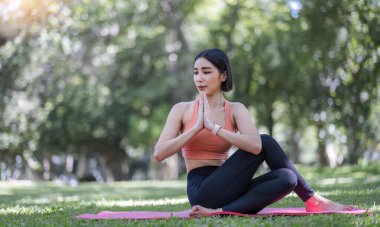 30 'lu yaşlarındaki çekici ve çekici Asyalı kadın spor kıyafetleriyle yoga yapıyor, kaslarını gevşetiyor. Güzel yeşil bir parkta yoga yapan Asyalı kadın..