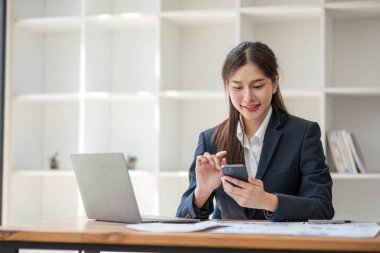 İnternetteki toplantıda dizüstü bilgisayar kullanan çekici Asyalı kadın. İş kadını cep telefonu kullanıyor. Modern ofiste masada oturan iş arkadaşıyla konuşuyor..