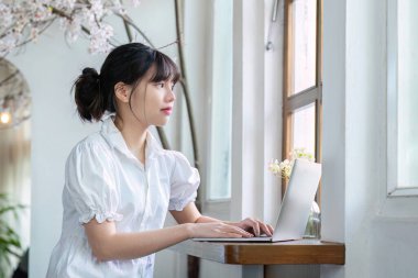 Oturma odasında dizüstü bilgisayarla çalışan Asyalı bir kız öğrenci bir ders için online araştırma yapıyor kompozisyon ödevi için notlar alın. çevrimiçi eğitim kavramı.