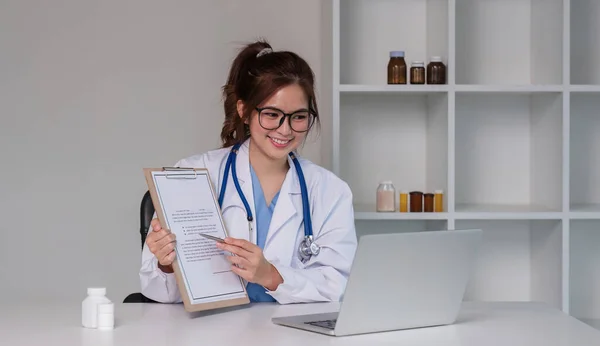 young asian lady doctor in white medical uniform with stethoscope using computer laptop talking video conference call with patient at desk in health clinic