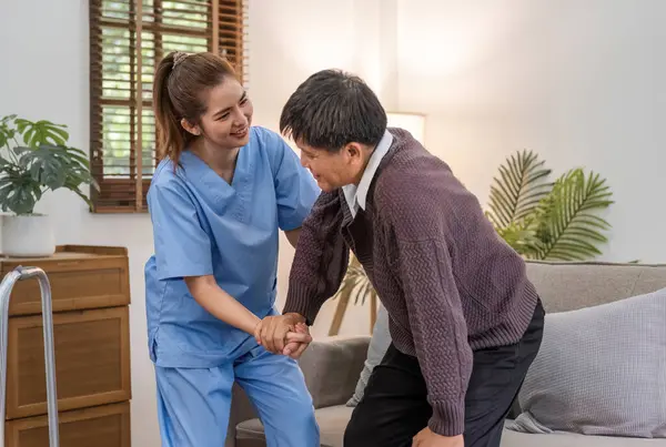 A caring young Asian nurse helps an elderly person walk in the house. An elderly disabled man tries to walk with the help of a female caregiver..