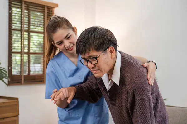 A caring young Asian nurse helps an elderly person walk in the house. An elderly disabled man tries to walk with the help of a female caregiver..