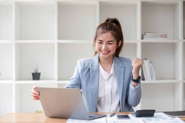 Asyalı iş kadınları, modern bir ofiste dizüstü bilgisayarla mutlu bir şekilde gülümseyen iş yerinde neşeli bir başarı ifadesi sergilerler...