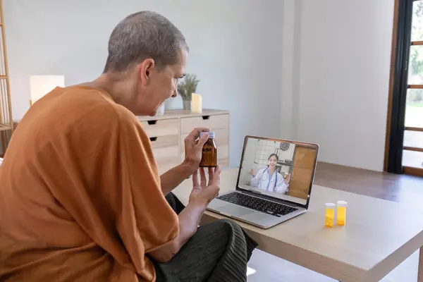 Elderly Woman 60s during Online Consultation with Doctor. Senior Female Patient Holding Bottle of Medicine while Discussing Medication with Health Care Provider over Internet at Home..