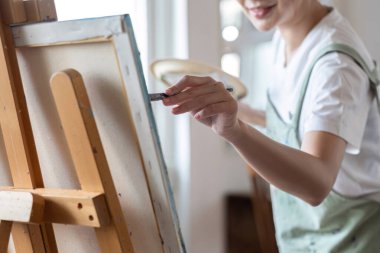 Yakından, çekici genç Asyalı kadın resim stüdyosunda akrilik boyalarla resim yapmaya odaklanıyor. Neşeli bir gülümsemeyle.