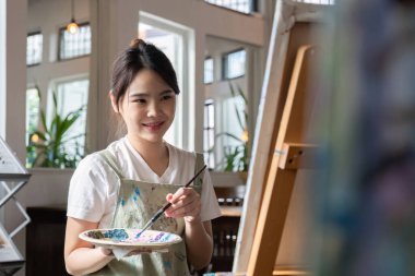 Çekici genç Asyalı kadın resim stüdyosunda akrilik boyalarla resim yapmaya odaklanıyor. Neşeli bir gülümsemeyle.