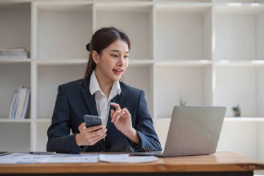 İnternetteki toplantıda dizüstü bilgisayar kullanan çekici Asyalı kadın. İş kadını cep telefonu kullanıyor. Modern ofiste masada oturan iş arkadaşıyla konuşuyor..