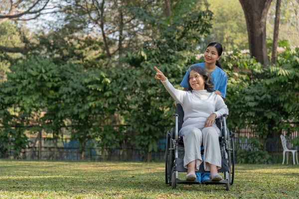 Happy nurse and elderly woman sitting in wheelchair enjoying outdoor. Beautiful nurse with senior woman in wheelchair at outdoor park. Smiling disabled old lady in wheelchair at park..