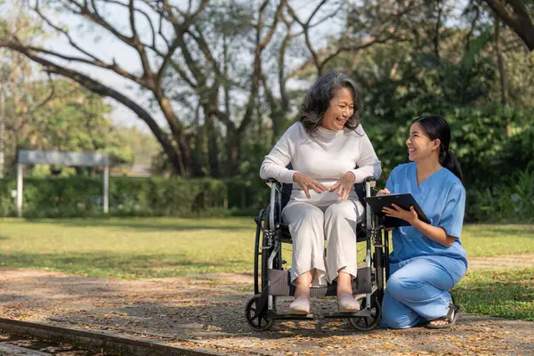Asian nurse or physiotherapist caring for elderly woman sitting wheelchair. Asian female nurse takes care of patients and takes them for a walk in the hospital park..