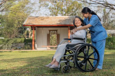 Üniformalı genç bir kadın ve tekerlekli sandalyedeki yaşlı bir hasta parkta takılıyor..