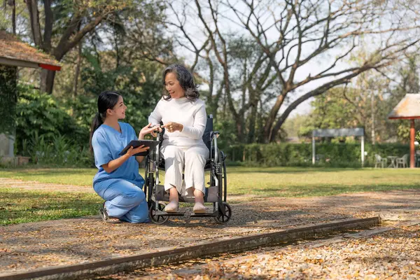 Asian nurse or physiotherapist caring for elderly woman sitting wheelchair. Asian female nurse takes care of patients and takes them for a walk in the hospital park..