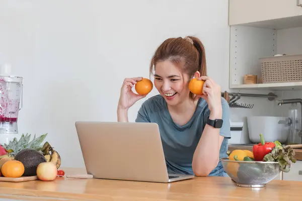 Beautiful young woman in exercise clothes having fun in a cute kitchen at home. Using your laptop to prepare vegan fruit salad dressing, fruit shakes, or healthy smoothies..