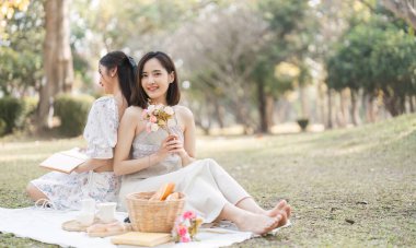 Gündelik giysiler ve yaz şapkaları içinde iki güzel LGBT 'li genç kadın dışarıda piknik yapıyor pozitif modelin önünde oturmuş meyve ve peynir yiyor ve selfie çekiyor. LGBT kavramı.