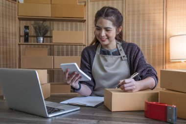 Genç bir Asyalı kadının portresi, arka planda paketlerle dolu ofiste oturan bir e-ticaret çalışanı KOBİ iş ekomünyesi ve teslimat işi için bir not ve hesap makinesi yazın...