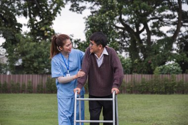 Asyalı kadın bakıcı, yaşlı bir adamın yürüteçle yürümesine yardım ediyor. Evde bir hemşire de ona yardım ediyor. Hemşire, huzurevindeki yaşlı bir hastaya yardım eder. Yaşlı bakımı ve sağlıklı yaşam tarzı.