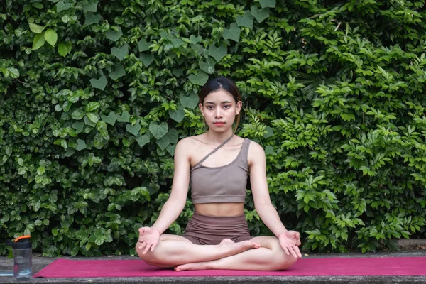 Young Asian woman doing yoga, meditation on a calm yoga mat in a green park in the early morning. Young woman wearing brown yoga clothes Yoga concept.