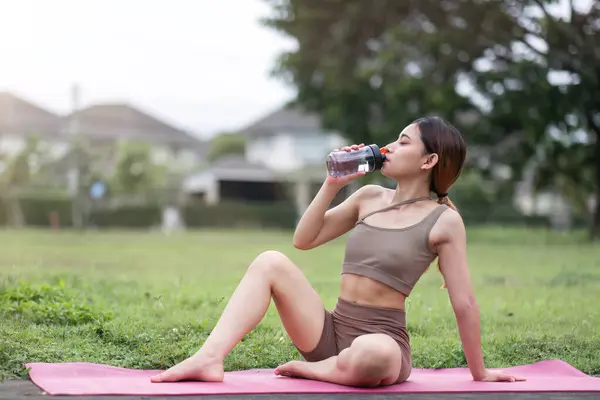 Beautiful young woman drink water after doing yoga in the village park. Yoga or fitness training health care concept.