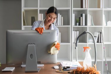 Genç bir kadın ofis masasındaki bilgisayar ve ekipmanları dezenfekte etmek için temizleme bezi kullanıyor. Temizlikçi ya da temizlikçi ofisi temizler..